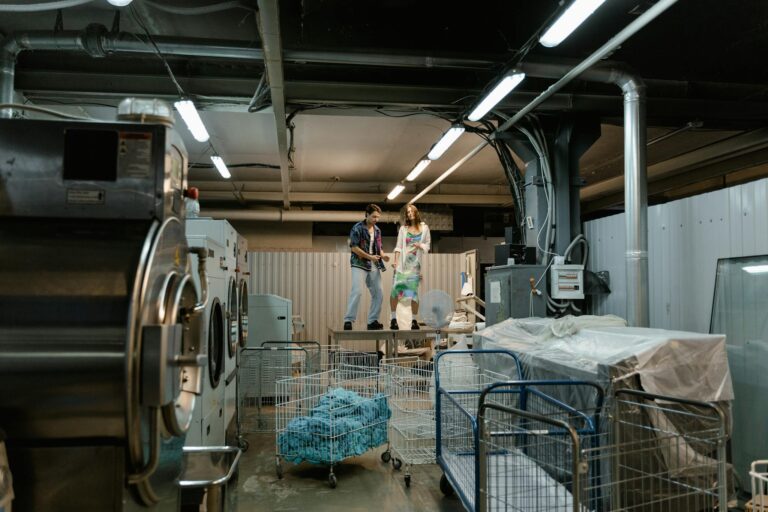 Two people working inside an industrial laundry facility with large machines and laundry carts.